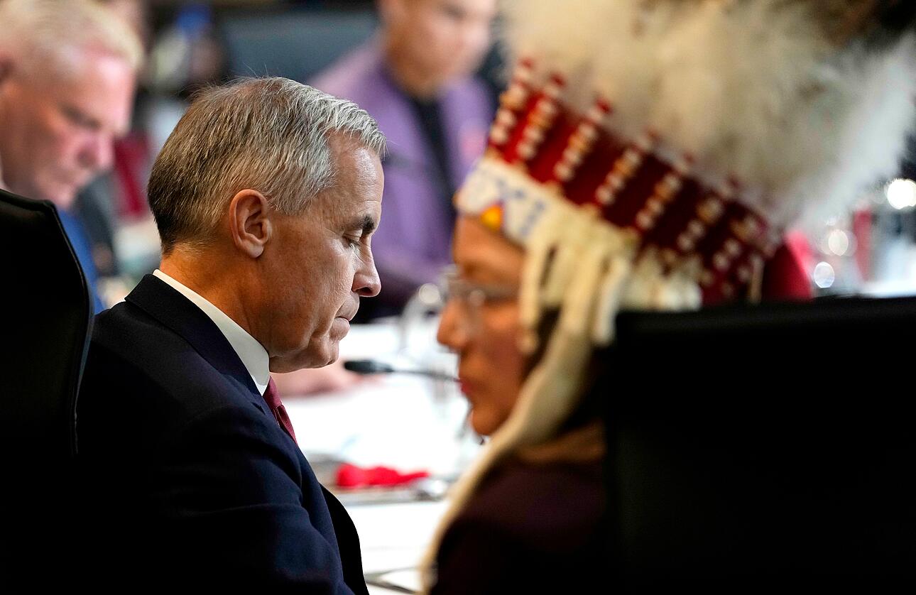 Prime Minister Mark Carney, in a dark suit, sits at a formal table with Indigenous leaders wearing traditional regalia with red and gold details in the background.